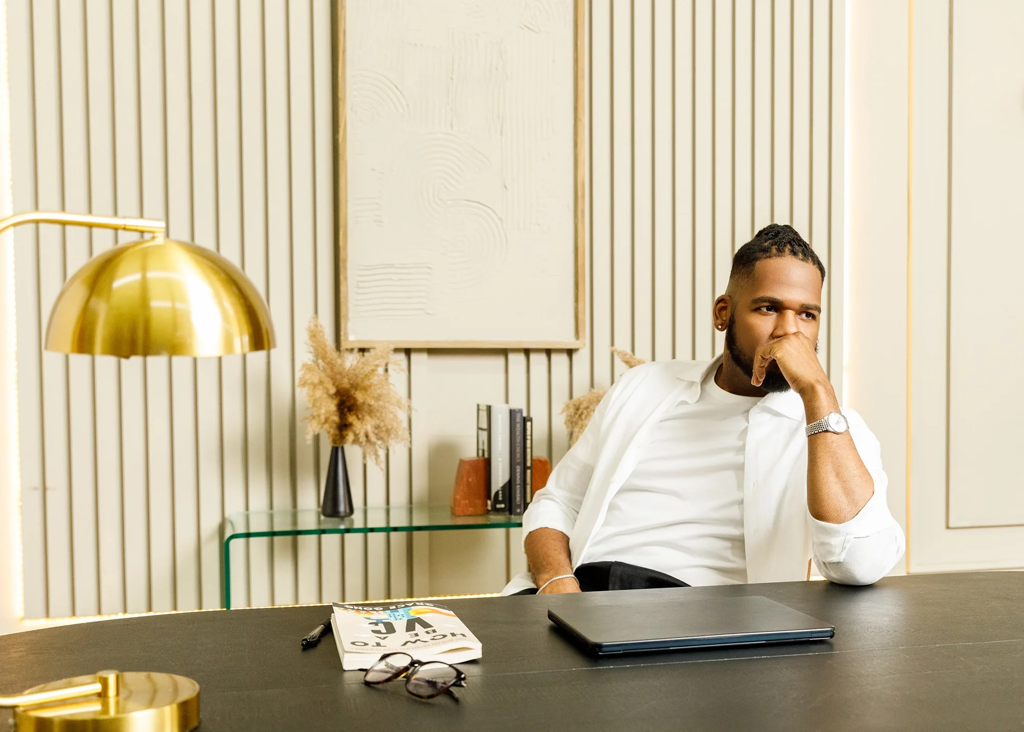 Brandon looking contemplative, leaning back in a chair at a desk with a lamp, book, and laptop on it