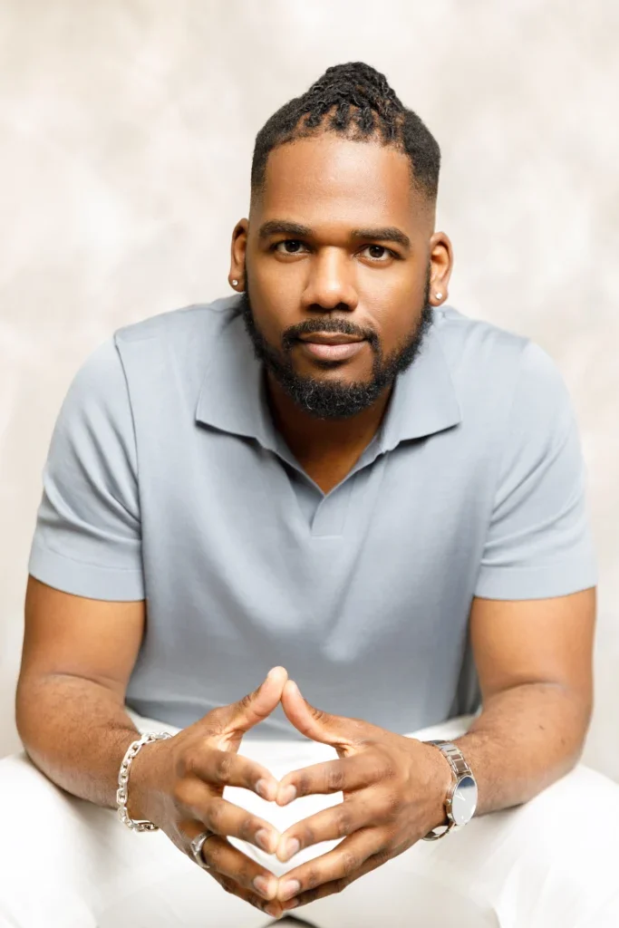 Brandon sitting again a beige backdrop with his hands together looking contemplative, wearing a light blue collared shirt