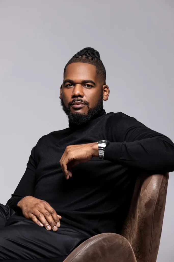 Brandon leaning back in a leather chair against a gray backdrop, he is wearing a black turtleneck sweater