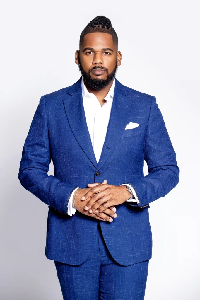Brandon against a gray backdrop wearing a blue suit with his hands clasped